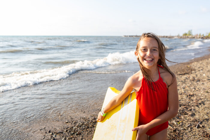 A girl with a body board smiling to the camera with Lake Erie in the background